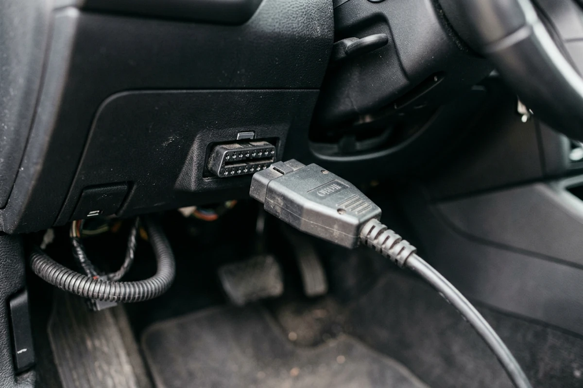 Handheld OBD-II scanner with a blank screen plugged into the diagnostic port under a car steering column